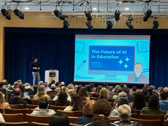 A speaker presenting to a large audience at a conference about 'The Future of AI in Education', with a presentation displayed on a screen.